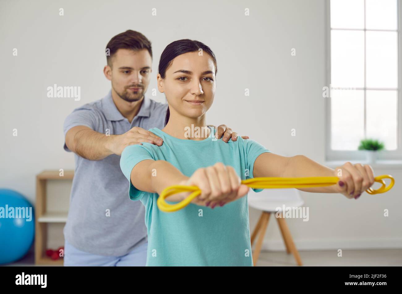 Medico fisioterapista maschile in uniforme aiuta ad esercitare le braccia con gomma elastica gialla alla giovane donna alla clinica di Riabilitazione. Foto Stock