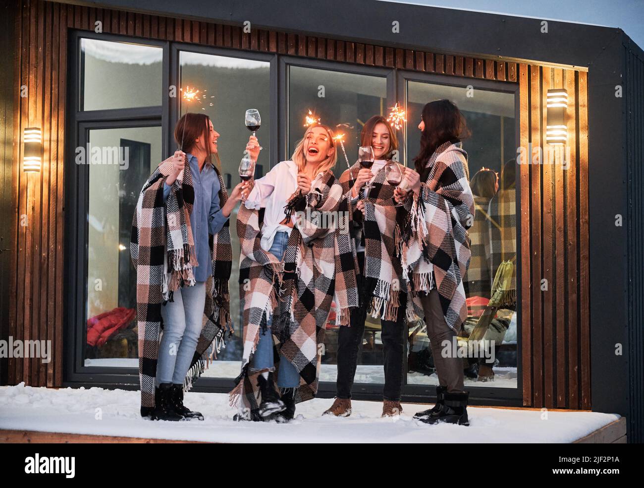 Le giovani donne che si divertiscono nei fine settimana invernali sulla terrazza della caserma contemporanea. Quattro ragazze in plaid bere vino e festeggiare con scintille la sera. Foto Stock