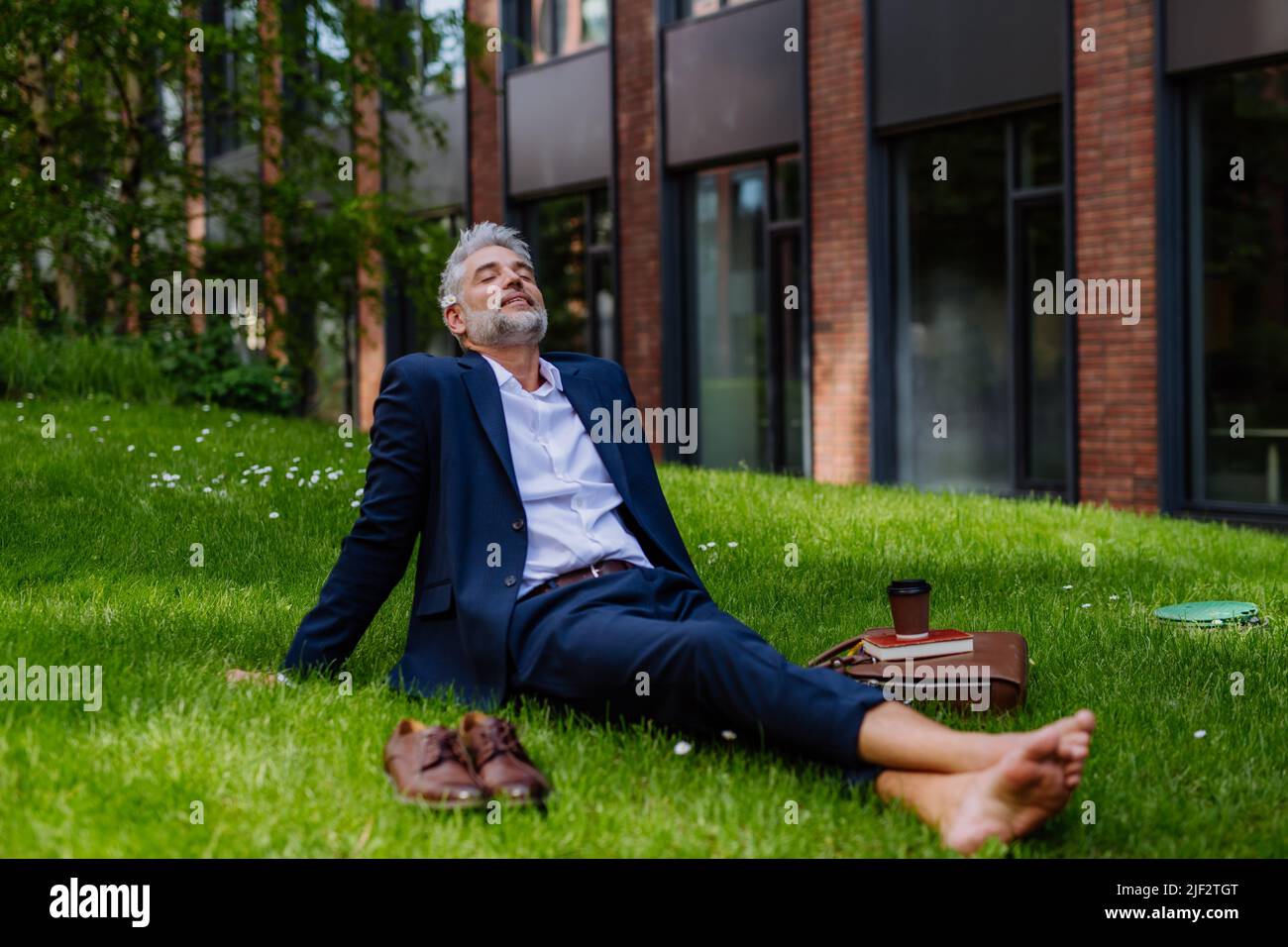 Uomo d'affari maturo che riposa e siede a piedi nudi nel parco, sentendosi libero, fuggendo dal lavoro, concetto di equilibrio di vita di lavoro. Foto Stock