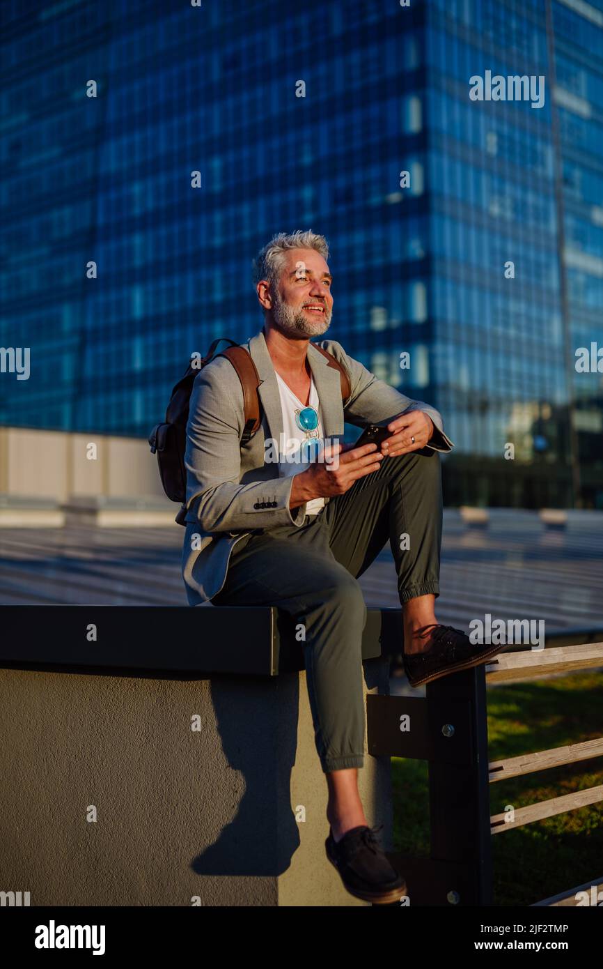 Uomo d'affari sicuro con zaino seduto sulle scale e con il telefono, sentirsi liberi, fuggire dal lavoro, concetto di equilibrio della vita lavorativa. Foto Stock