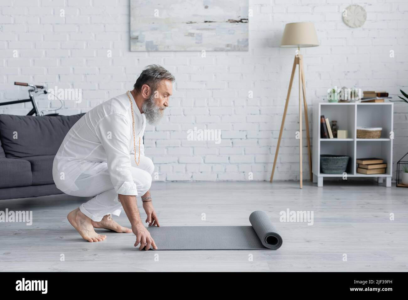vista laterale dell'uomo anziano in abiti bianchi che srotolano materassino yoga a casa Foto Stock