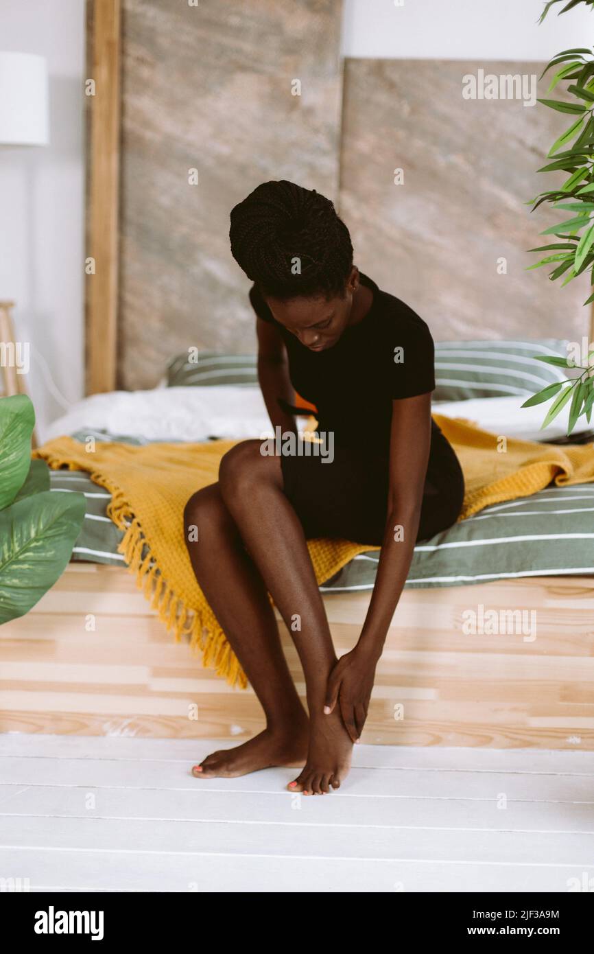 Bella donna afroamericana con dreadlocks seduta sul letto in studio decorato e massaggiante piede a mano. Persona nera che ha lesioni sportive, muscolo Foto Stock