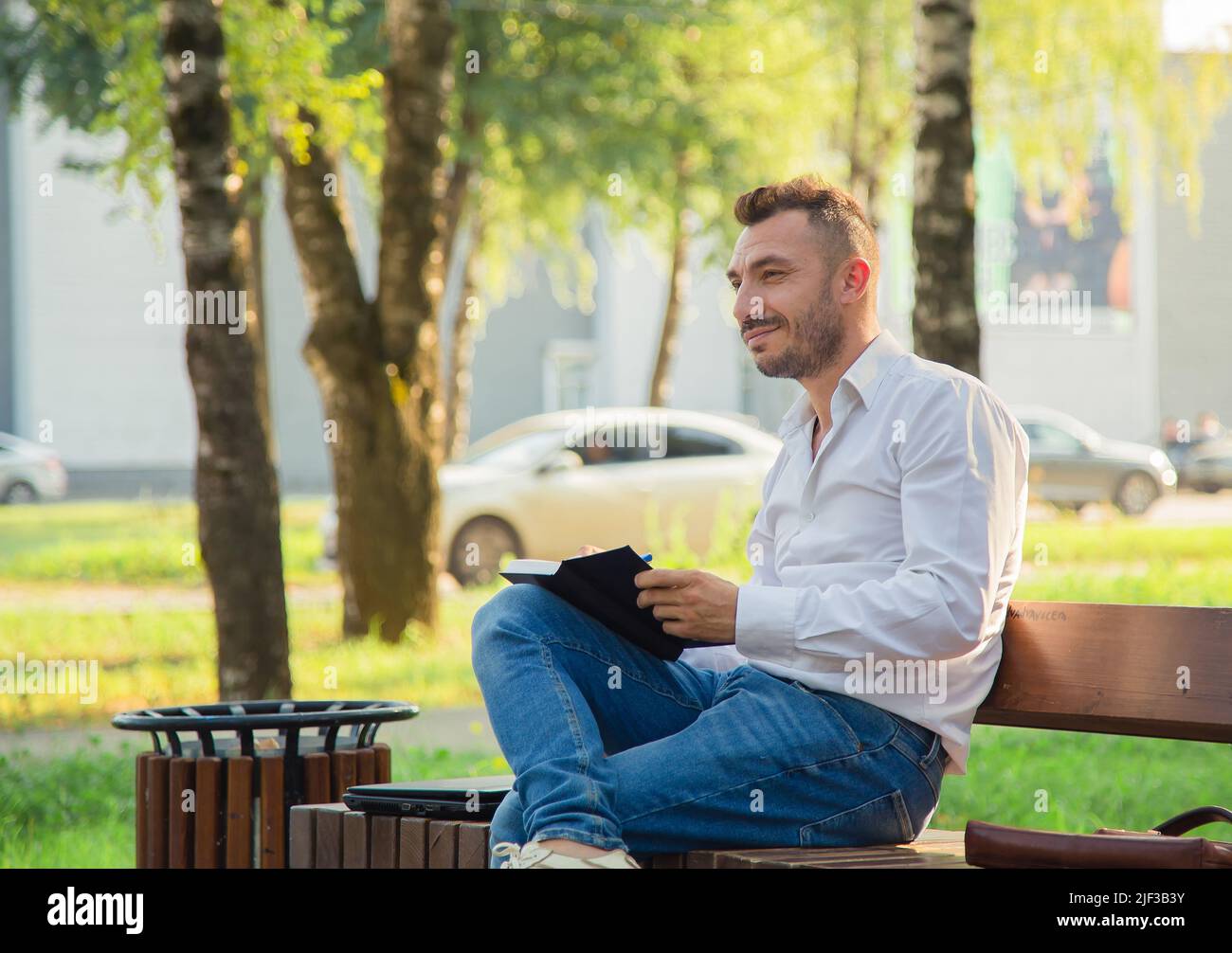 Un uomo felice si siede su una panca, fa i piani, scrive in un taccuino. Un giovane uomo su uno sfondo di alberi verdi, una calda giornata estiva di sole. Luce calda e soffusa, primo piano. Foto Stock