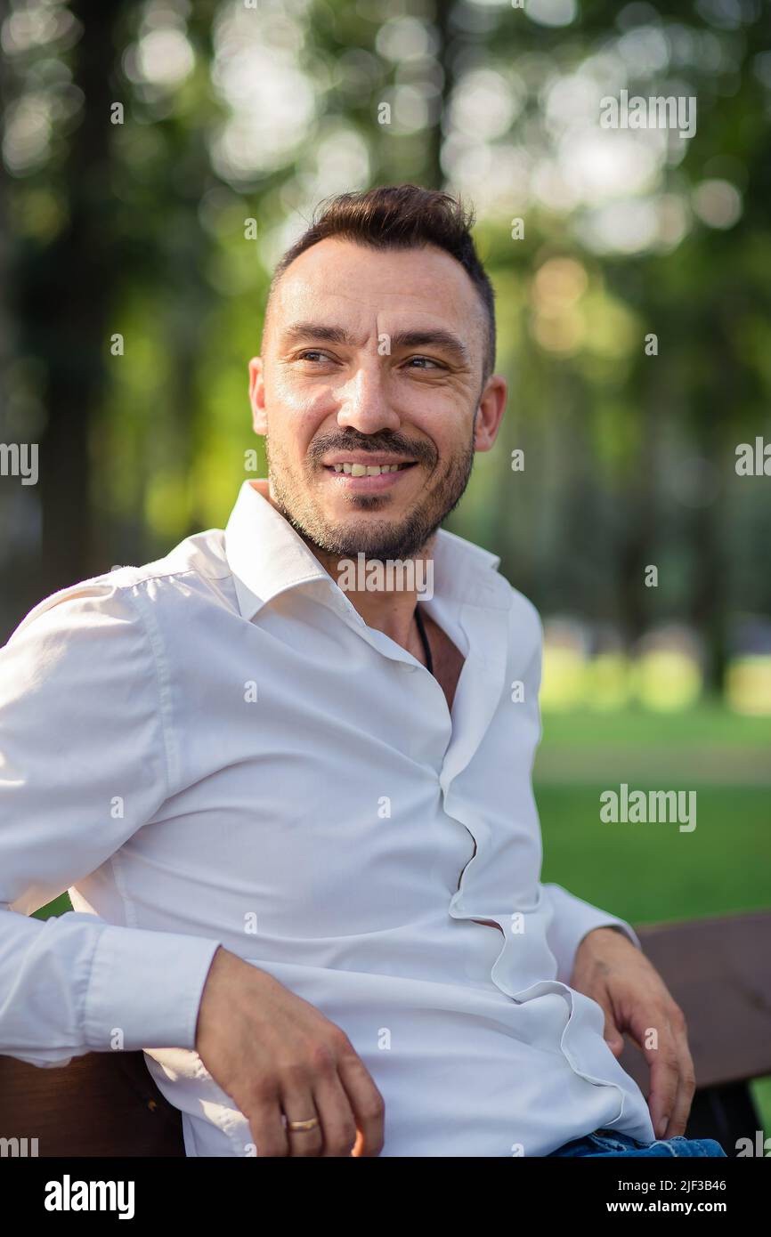 Sorridente bell'uomo in una camicia bianca è seduto su una panca. Giovane uomo nel parco della città, caldo giorno d'estate soleggiato. Luce calda e soffusa, primo piano. Foto Stock