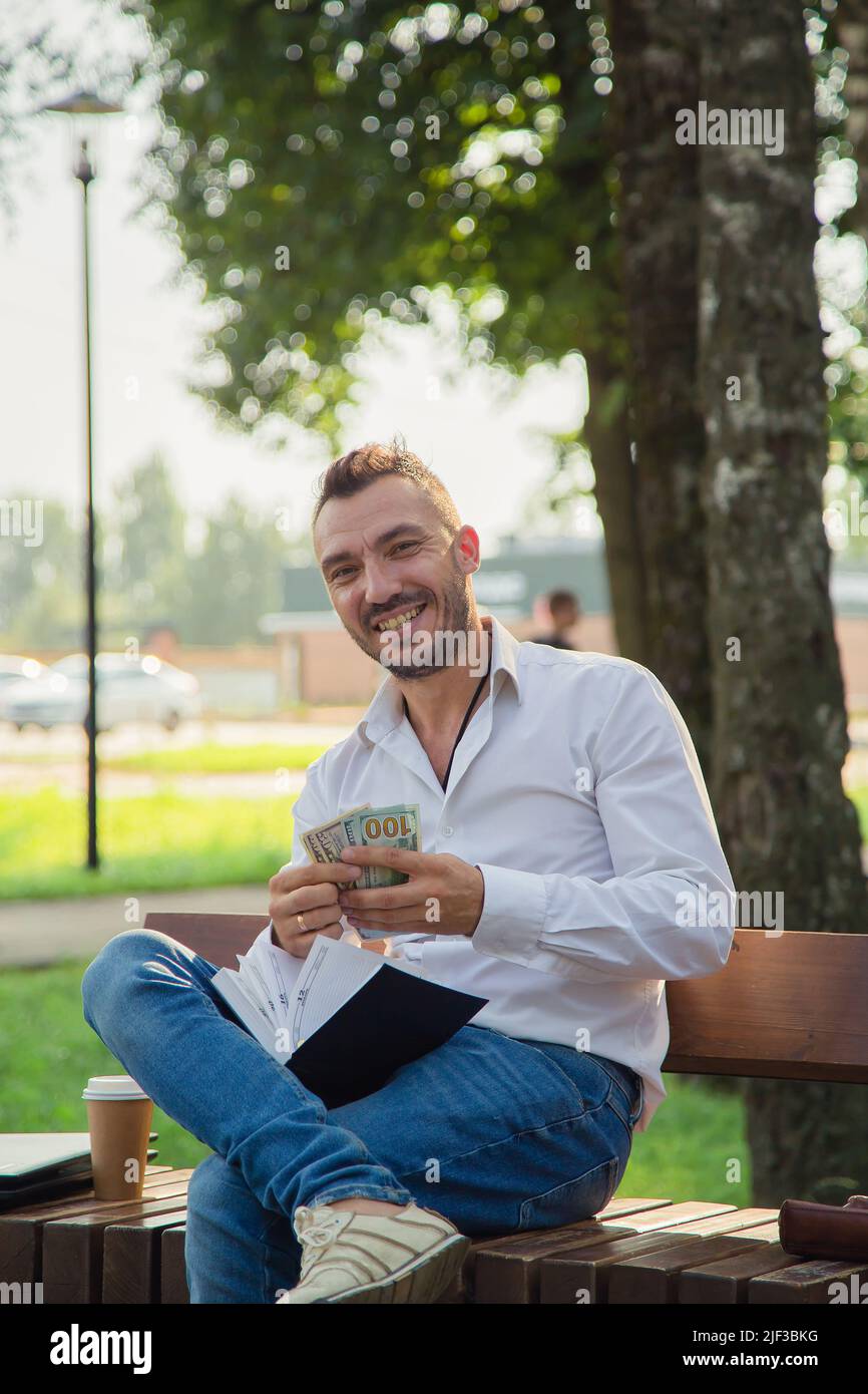 Un uomo felice è seduto su una panchina nel parco, contando dollari. Un giovane uomo su uno sfondo di alberi verdi, una calda giornata estiva di sole. Luce calda e soffusa, primo piano. Foto Stock
