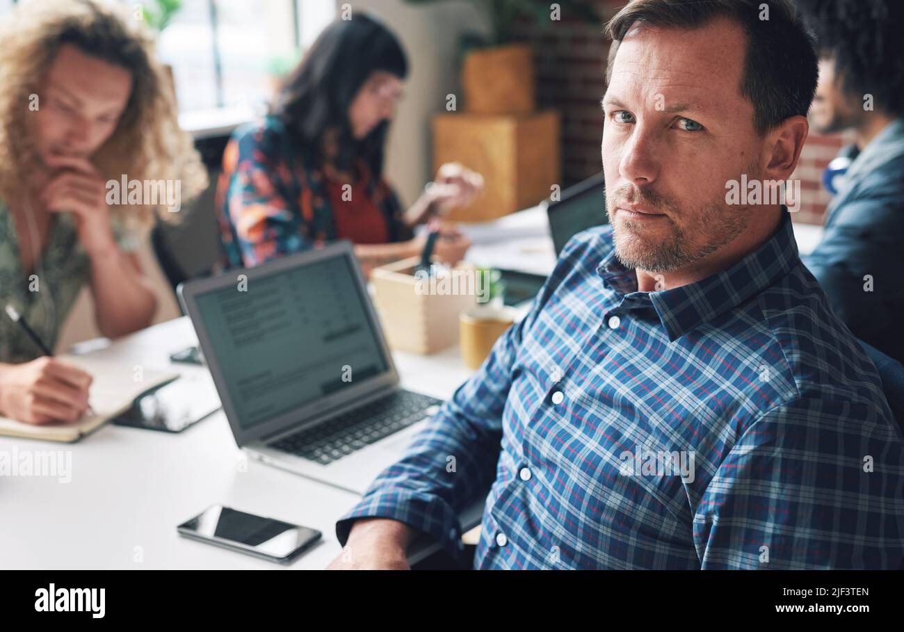 Ritratto di un maturo uomo d'affari serio seduto alla sua scrivania nel suo ufficio con un gruppo di colleghi diversi. Un uomo d'affari serio e leader nel suo Foto Stock