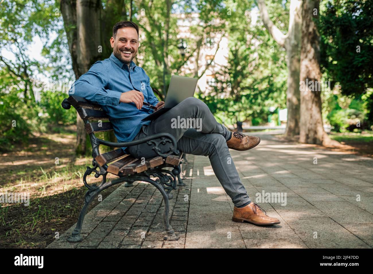 Ritratto di giovane imprenditore sorridente utilizzando il portatile. I formali professionali maschili sono seduti sulla panca. Lavora al parco della città. Foto Stock