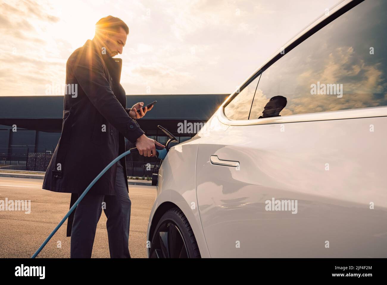 Un uomo d'affari elegante che carica una nuova auto elettrica bianca, nel parcheggio del centro commerciale al tramonto Foto Stock