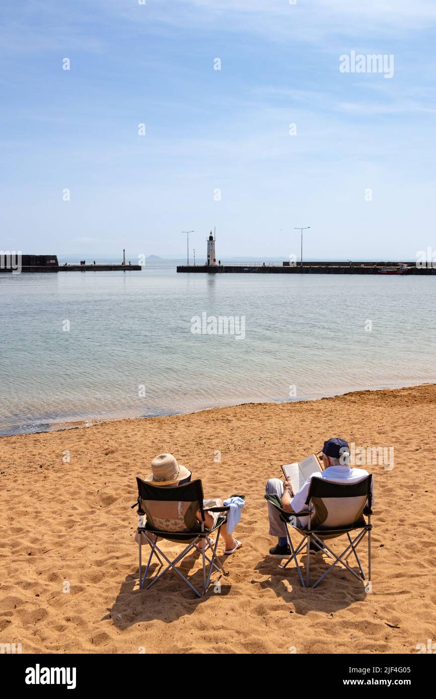 Due adulti anziani sedevano su sedie portatili su una spiaggia sabbiosa che si affaccia lontano, in direzione del muro del porto e del faro o dell'aiuto alla navigazione Foto Stock