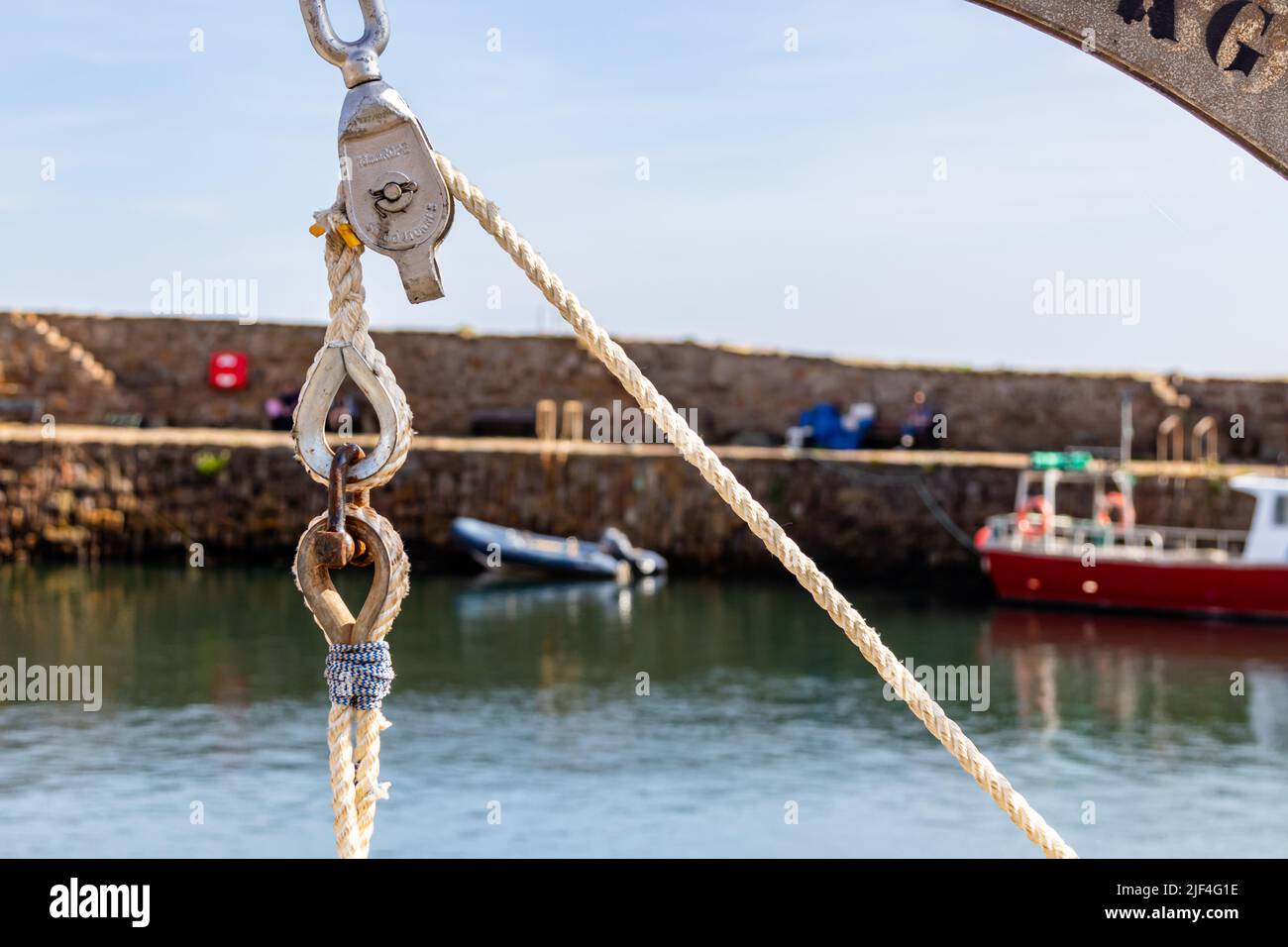 Primo piano di una fune, una puleggia e un gancio al sole, contro una parete del porto fuori fuoco o una banchina con le barche sullo sfondo, incluso un gommone Foto Stock