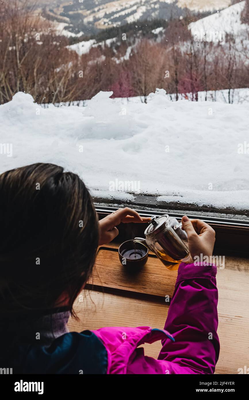 Donna che beve il tè vicino alla grande finestra con vista sulle montagne invernali. Atmosfera accogliente Foto Stock