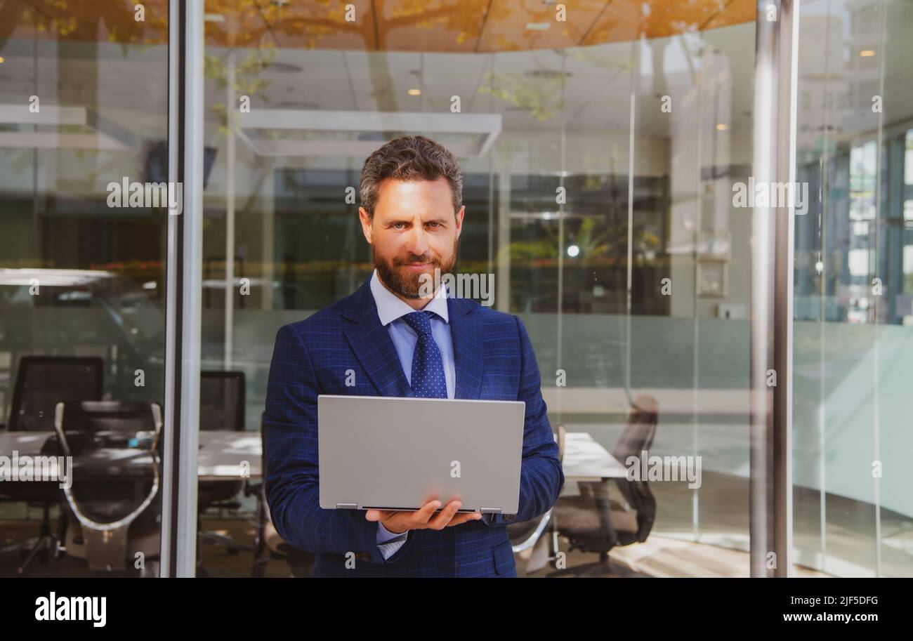 Uomo d'affari che tiene un computer portatile in piedi sulla strada vicino all'edificio degli uffici aziendali. Foto Stock