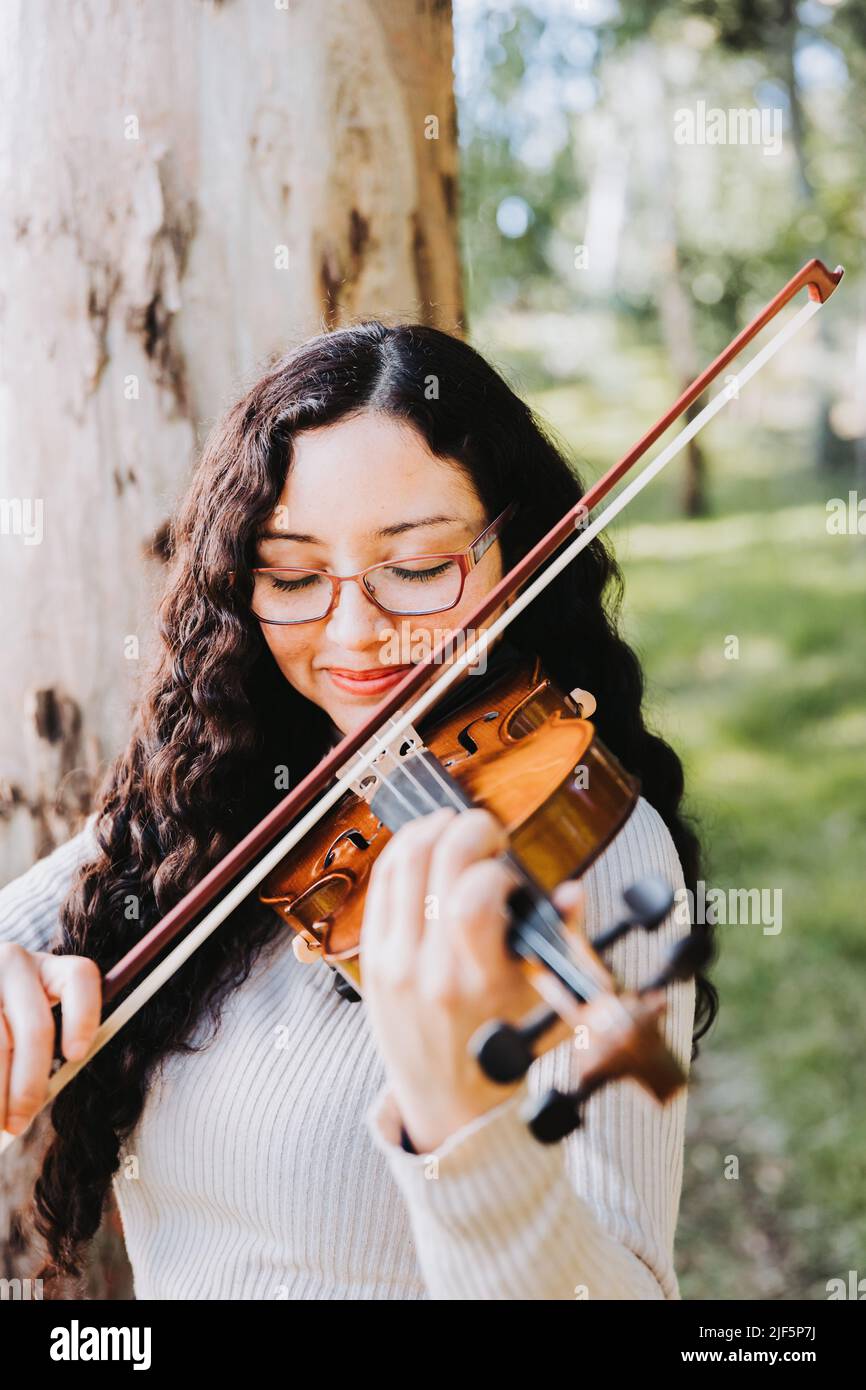 Donna bruna sorridente con occhiali che suonano violino all'esterno nei boschi. Verticale Foto Stock