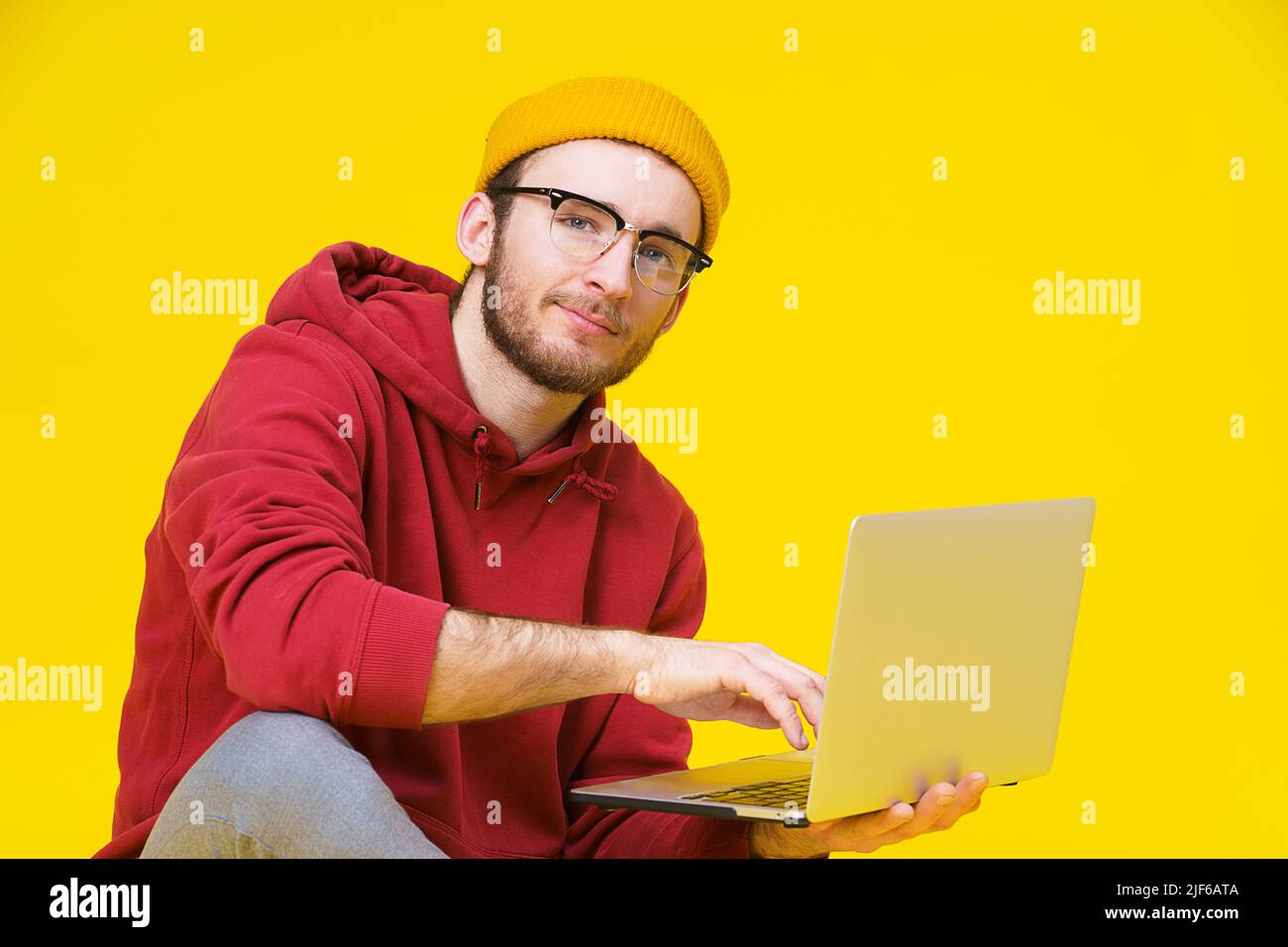 Giovane uomo hipster seduto sul pavimento che tiene un computer portatile con cappuccio rosso che studia o lavora. Uomo caucasico freelance intelligente con notebook isolato su sfondo giallo. Foto Stock