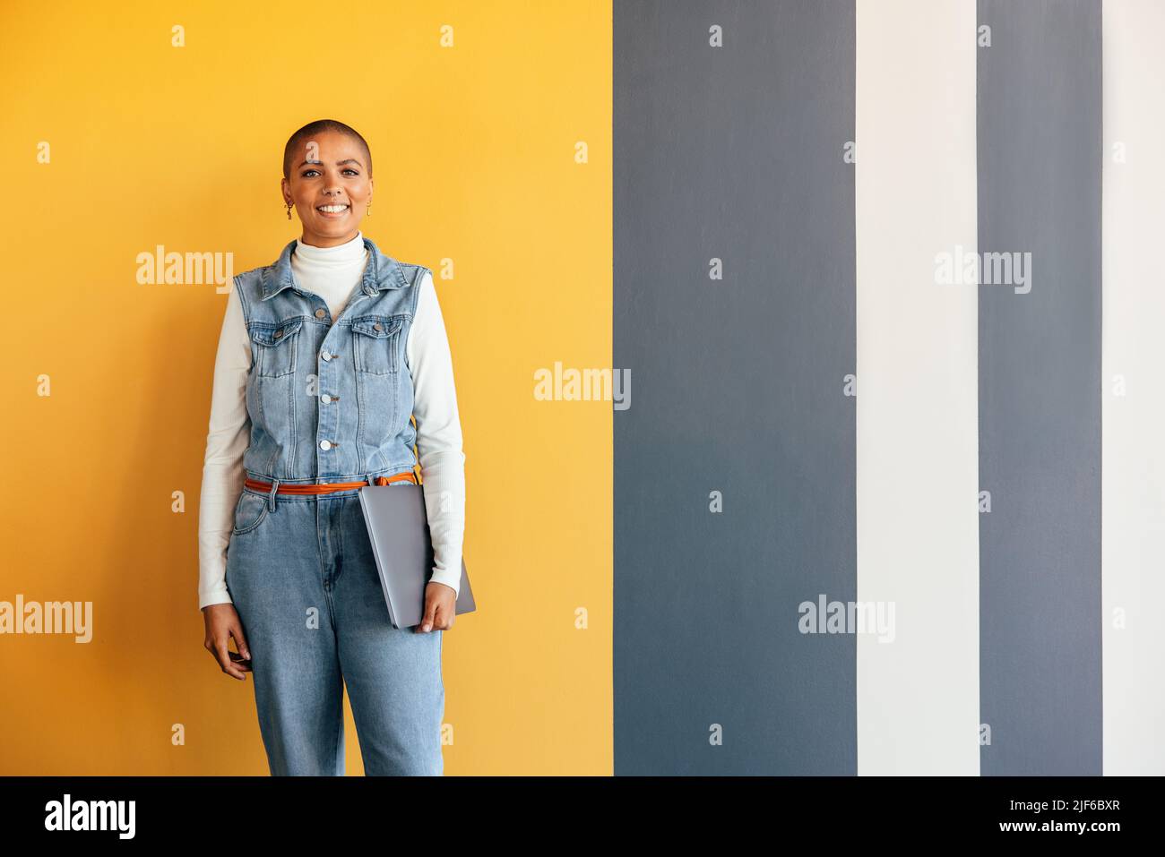 Donna d'affari moderna sorridente alla macchina fotografica mentre si alza in piedi da sola in un ufficio. Giovane donna d'affari allegra che tiene un laptop mentre si trova contro un c Foto Stock