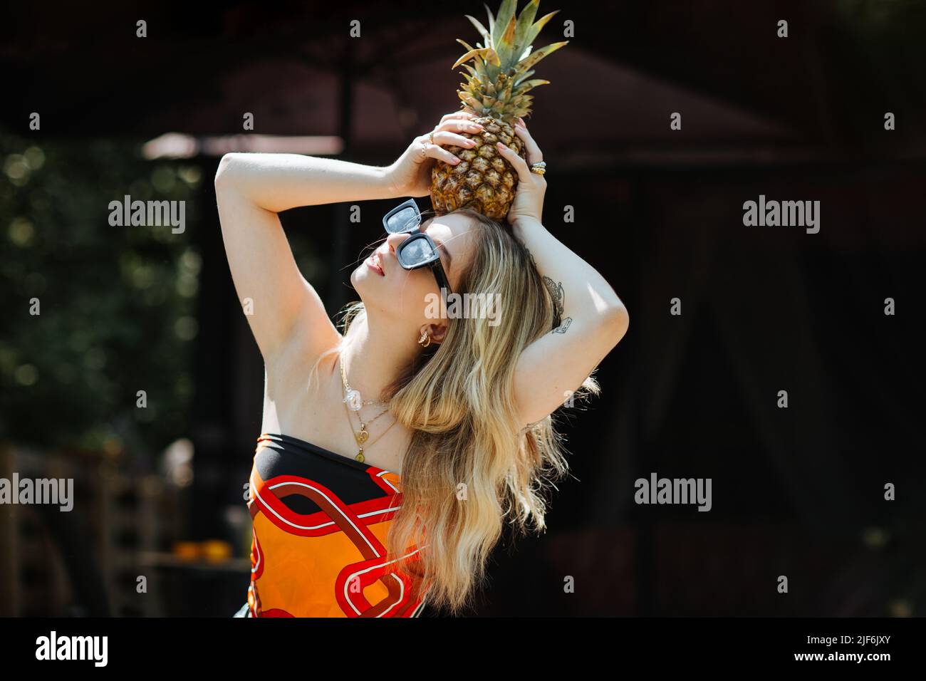 Giovane donna in elegante top e occhiali da sole con ananas maturo sulla testa, mentre in piedi sulla terrazza dell'edificio Foto Stock