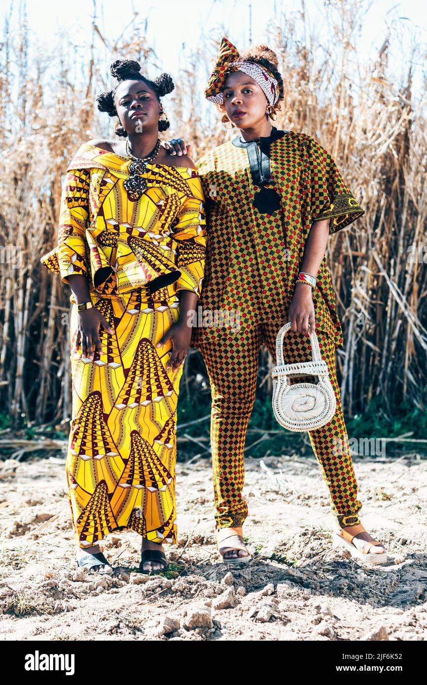 Modelli femminili neri sicuri in abiti eleganti con stampe africane autentiche che guardano la macchina fotografica mentre si levano in piedi insieme contro l'erba asciutta nel conteggio Foto Stock