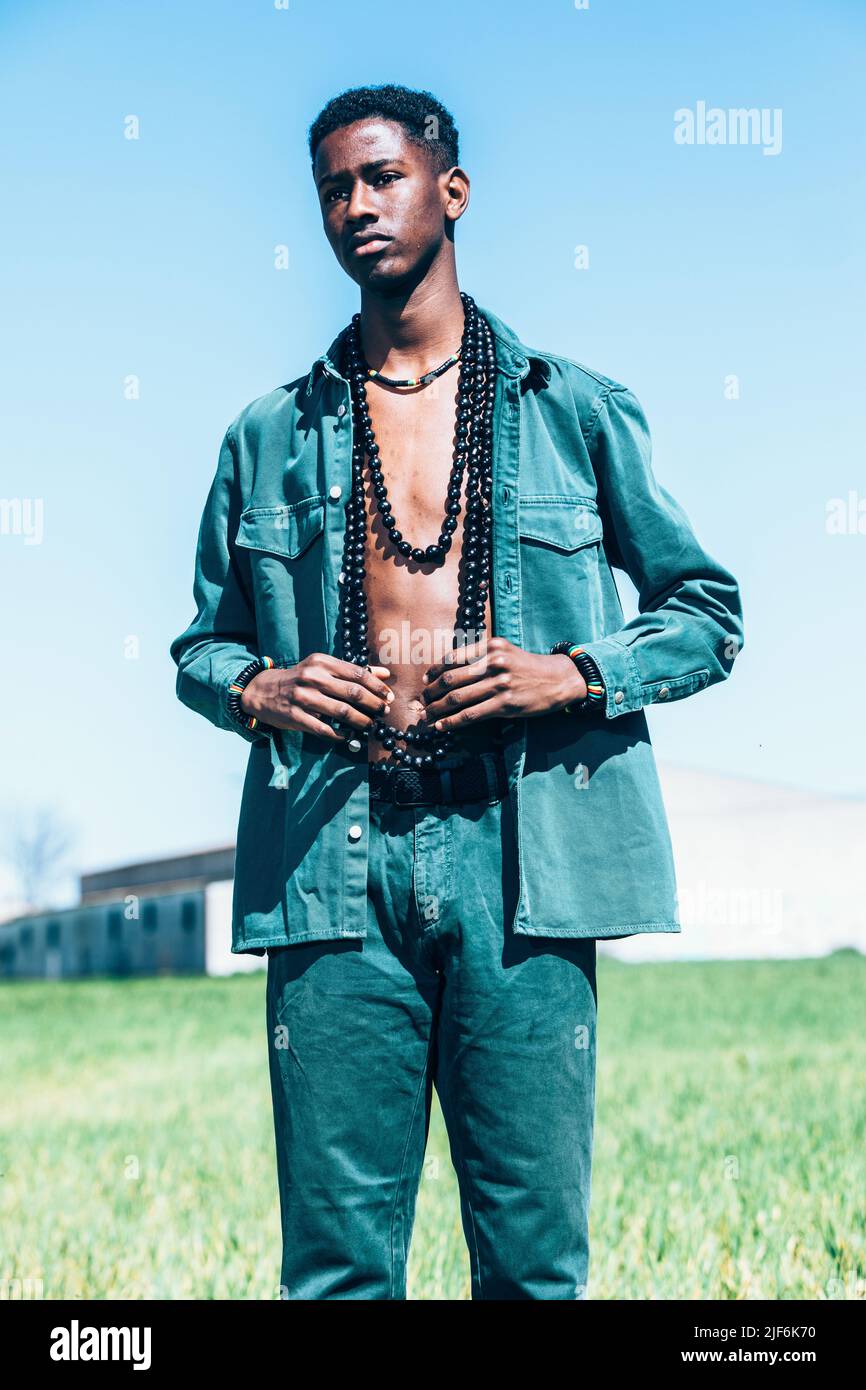 Giovane uomo nero in elegante abito in denim con collana tradizionale che guarda via mentre si alza in erba verde contro il cielo blu in terreno rurale Foto Stock