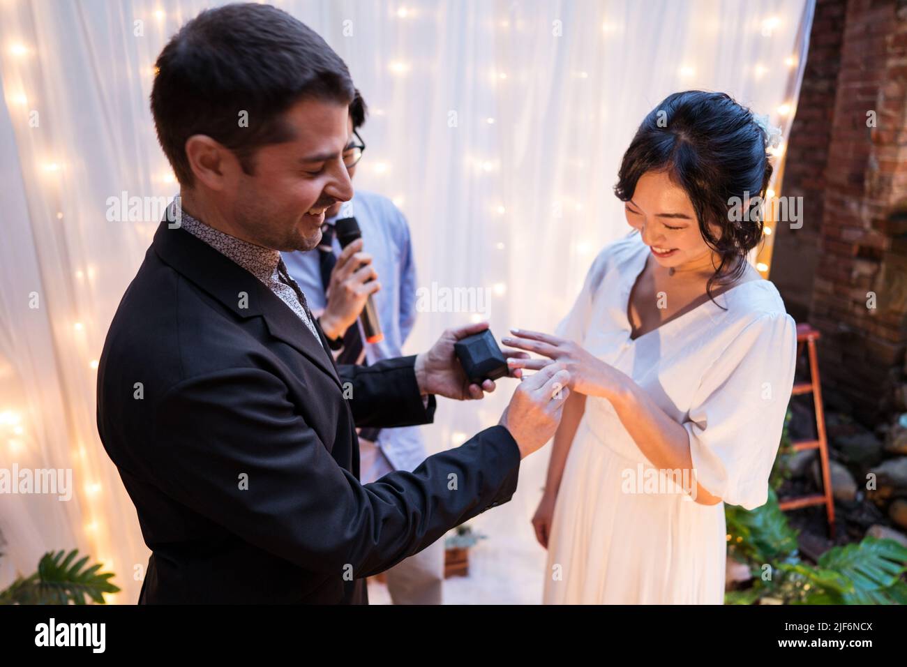 Alto angolo di coppia multietnica newlywed sorridente mentre si alza vicino arco decorato con cortina e bulbo chiaro ghirlande mentre si scambiano anelli durin Foto Stock