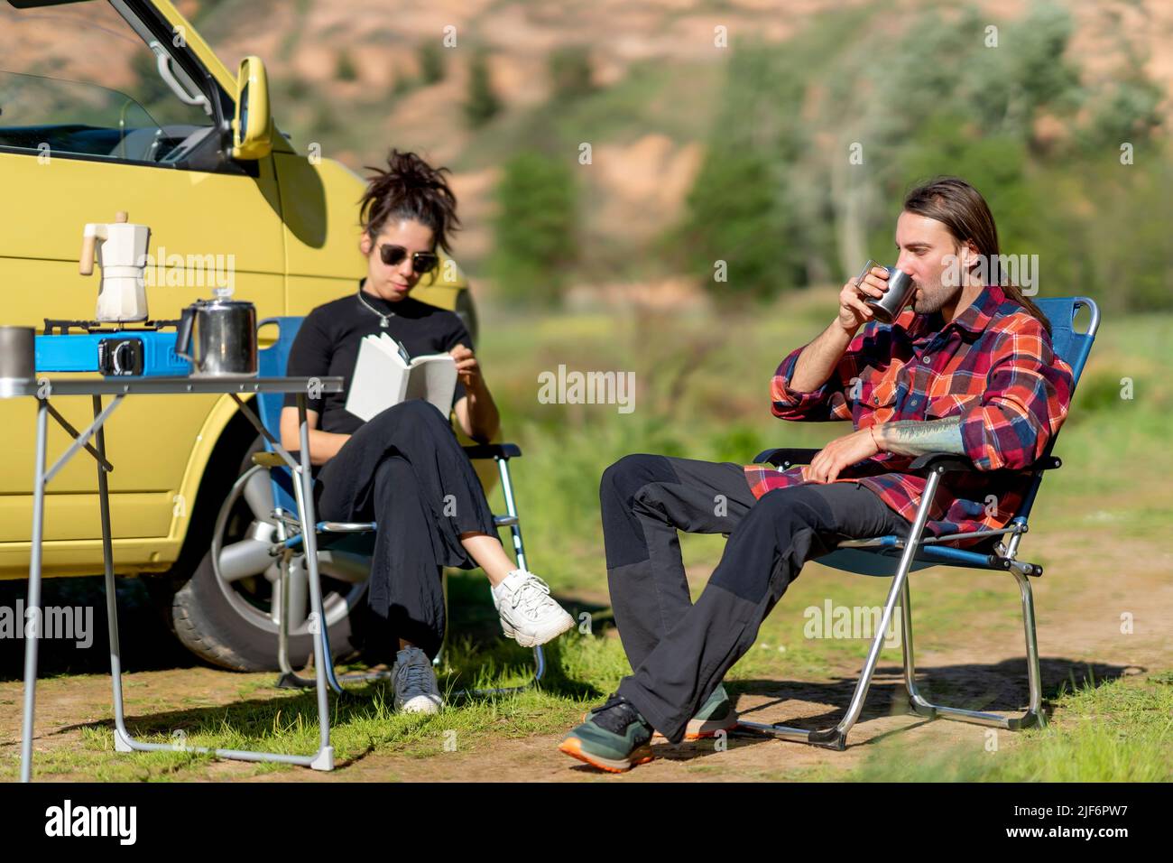 Corpo completo Ispanica Donna in abiti casual e occhiali da sole leggere libro mentre si siede sulla sedia vicino all'uomo in camicia a scacchi bere caffè in campeggio Foto Stock