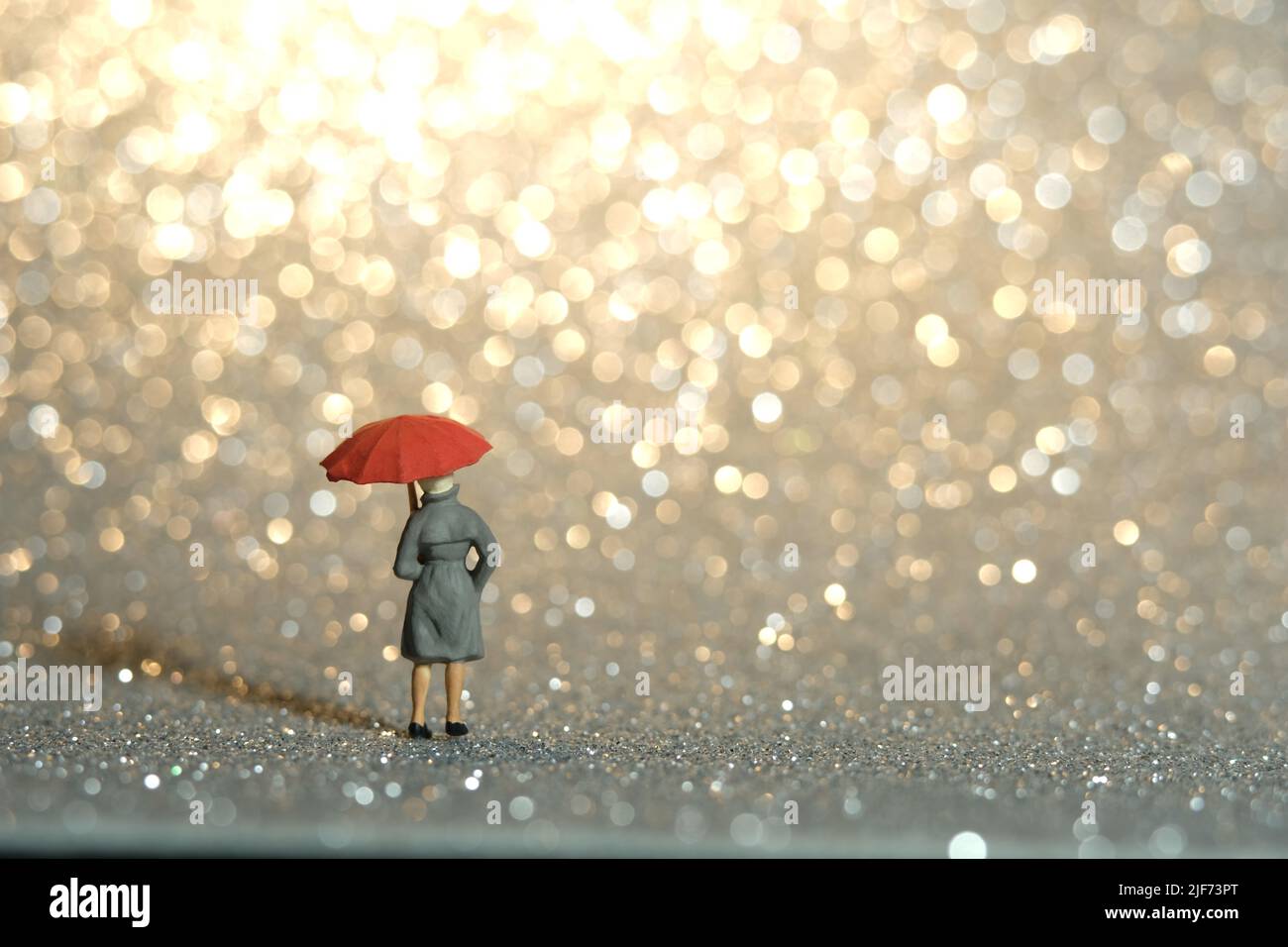 Fotografia di personaggi giocattolo in miniatura. Una signora che indossa impermeabile e ombrello, in piedi godendo di oro giallo bokeh sfondo chiaro. Foto immagine Foto Stock
