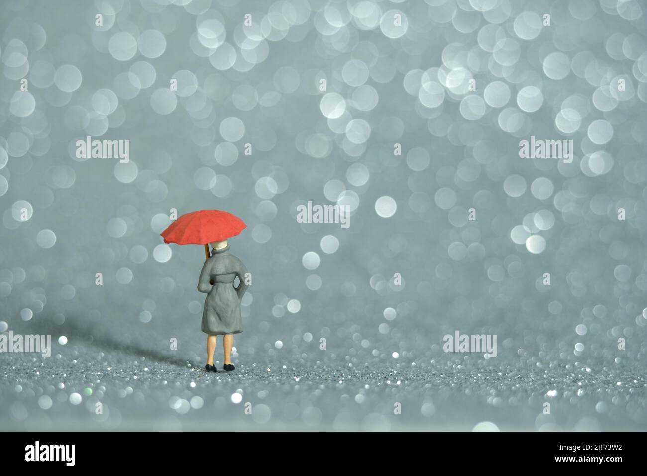 Fotografia di personaggi giocattolo in miniatura. Una signora che indossa impermeabile e ombrello, in piedi godendo di bianco argento bokeh sfondo chiaro. Foto immagine Foto Stock