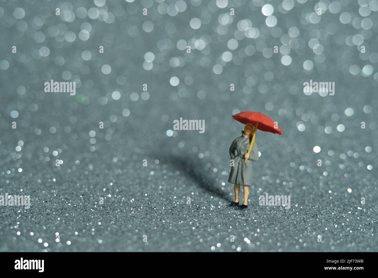 Fotografia di personaggi giocattolo in miniatura. Una signora che indossa impermeabile e ombrello, in piedi godendo di bianco argento bokeh sfondo chiaro. Foto immagine Foto Stock