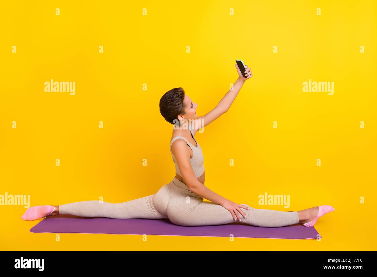 Profilo laterale ritratto di attraente ragazza seduta su tappeto allenarsi spago facendo selfie isolato su sfondo giallo brillante Foto Stock