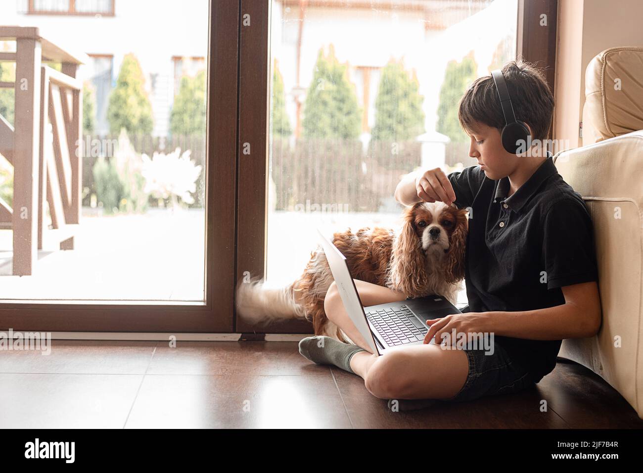 Vista laterale del ragazzo adolescente seduto sul pavimento con il cane Cavalier re Charles spaniel alla porta del patio, stroking, alimentazione animale domestico. Foto Stock