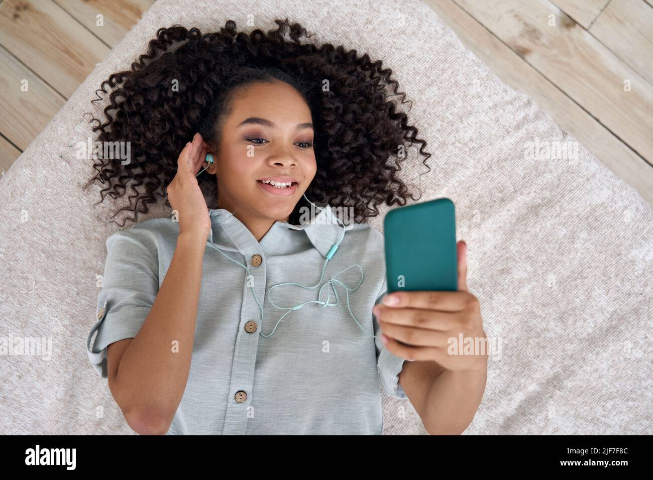 Felice ragazza africana teen in auricolari sdraiati a letto ascoltare musica su smartphone. Foto Stock