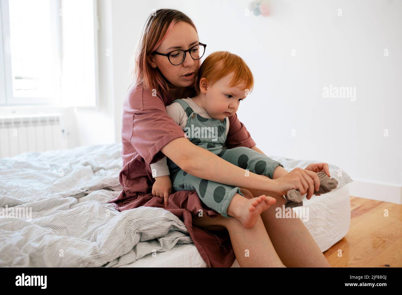 La madre mette calzino al piede del toddler maschio che si siede in camera da letto Foto Stock