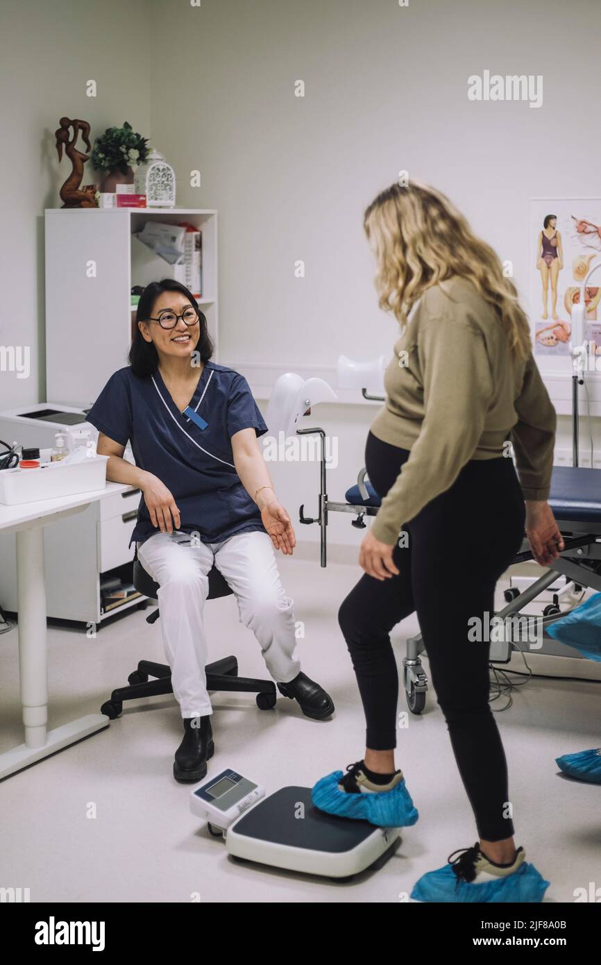 Tutta la lunghezza del medico sorridente che guarda la donna incinta in piedi sulla scala di peso in clinica medica Foto Stock