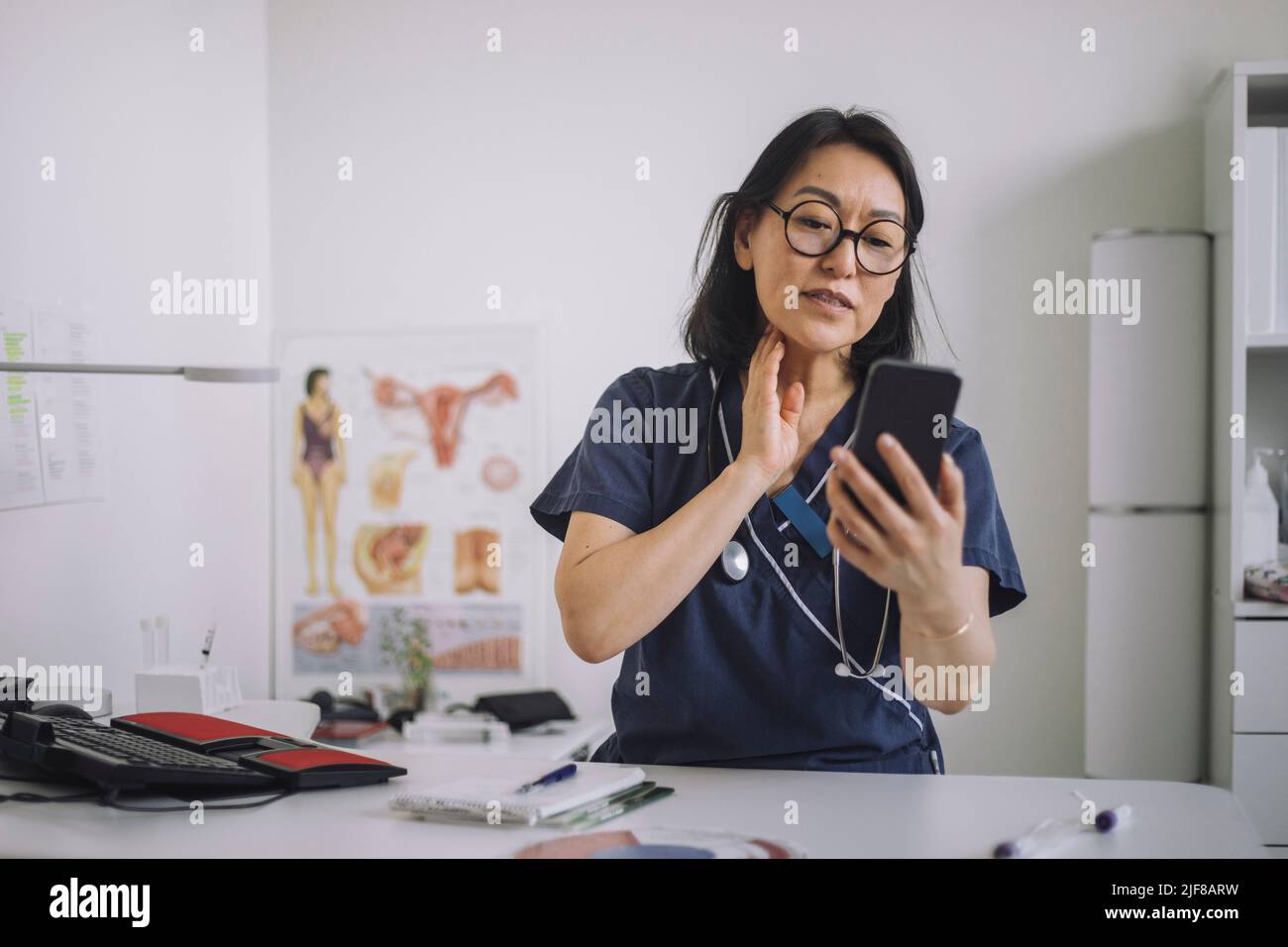 Otolaringologo femminile che fa la consultazione online attraverso il telefono cellulare mentre si siede alla scrivania in clinica medica Foto Stock