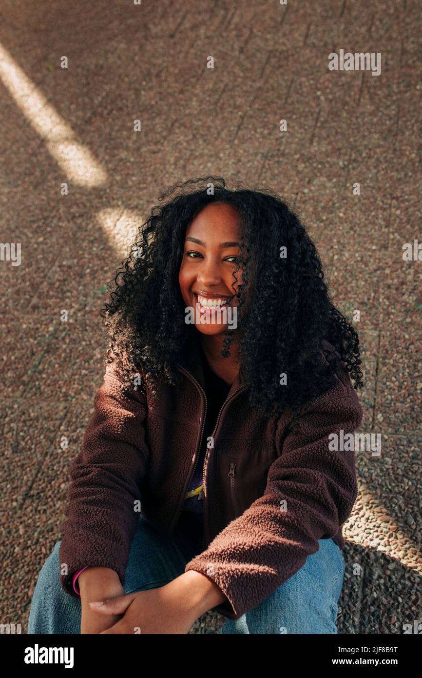 Ritratto di giovane donna sorridente con capelli ricci seduti a lato della strada Foto Stock