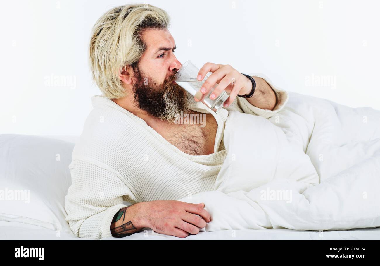 Uomo bearded in accappatoio bere acqua nel letto. Mattina e sveglia. Acqua. Rimani idratato. Liquido sano. Foto Stock