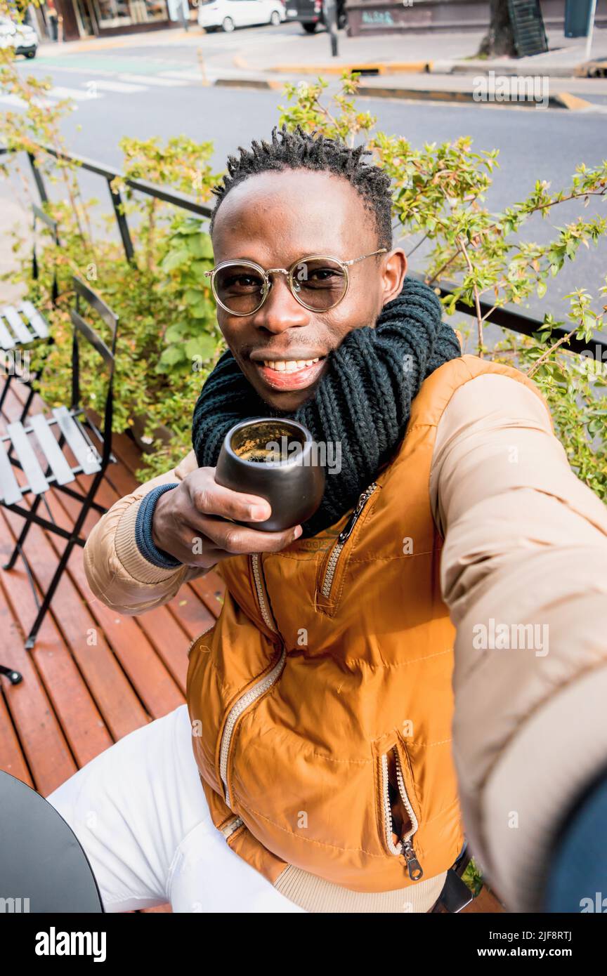 il giovane uomo africano che indossa occhiali, sciarpa e abiti invernali, fa selfie con il suo telefono bere caffè seduto al di fuori del bar, guardando camer Foto Stock