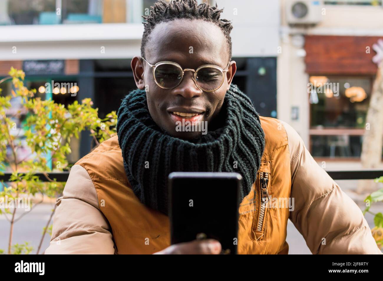 happy african uomo solo all'aperto, sorridere e controllare le notifiche e i messaggi dai social network e la posta sul suo telefono, in attesa di colazione, s Foto Stock