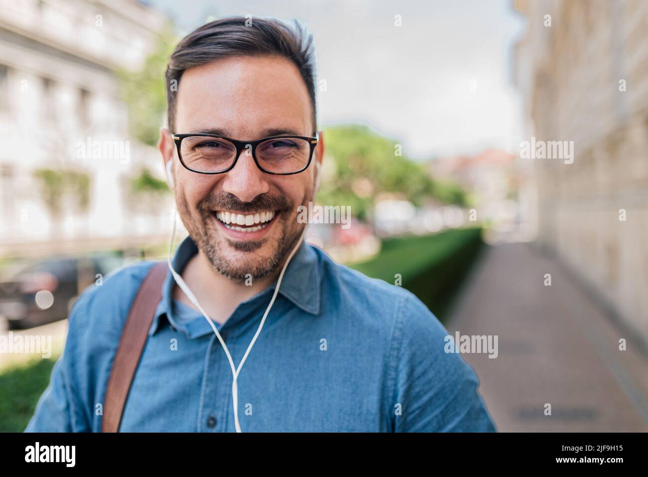Sorridente giovane uomo d'affari con le cuffie. Ritratto di maschio professionista commuter ascolto musica. Indossa occhiali e formals in città. Foto Stock