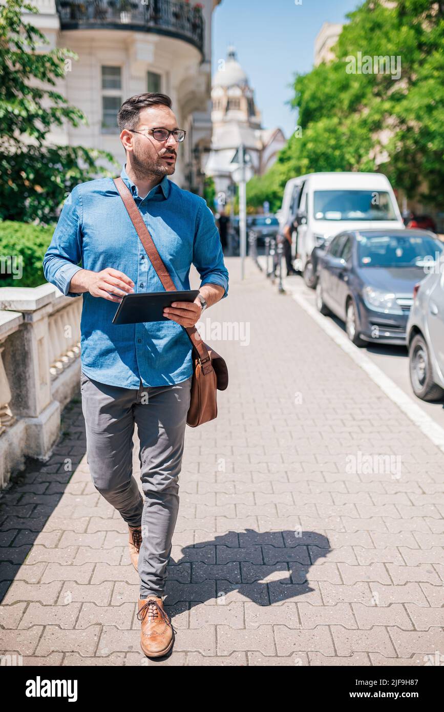 Un uomo d'affari sicuro con un tablet digitale che guarda via. Il pendolari professionista maschile cammina con i veicoli sul marciapiede. Indossa i formals in città d Foto Stock