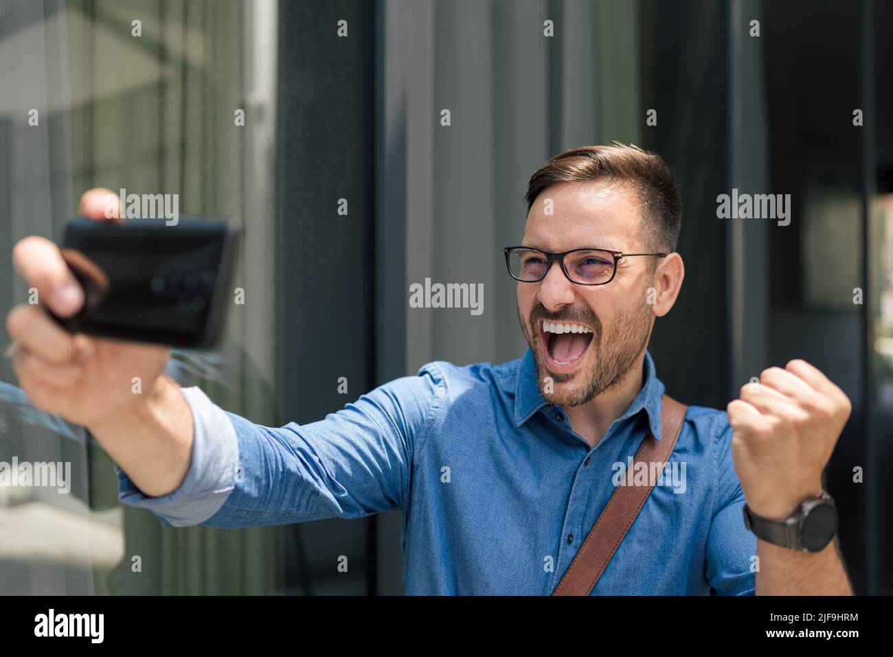 Felice uomo d'affari che prende selfie su smartphone. Il dirigente maschile sta aggrappando il pugno mentre gridava. Si trova in piedi contro l'edificio degli uffici della città. Foto Stock