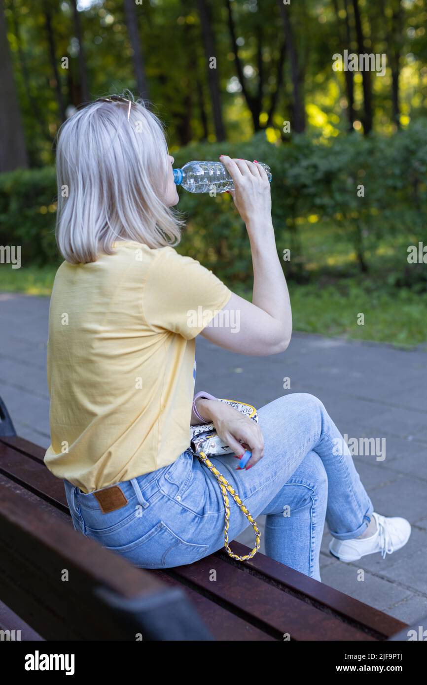 una ragazza siede su una panca nel parco e beve acqua da una bottiglia. Foto di alta qualità Foto Stock