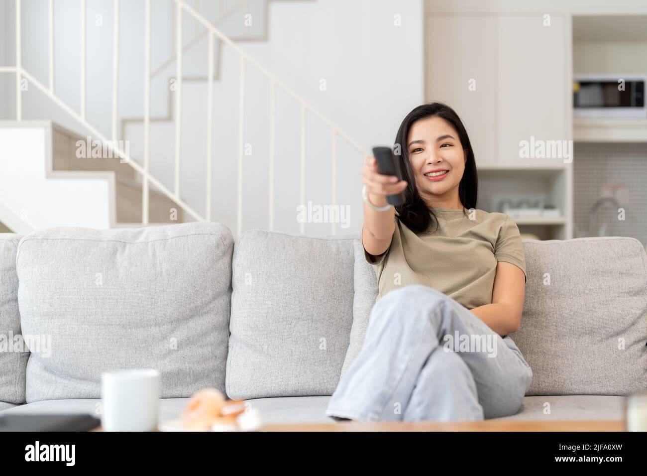 Immagine di una bella donna asiatica canale di ricerca con telecomando per guardare la tv mentre si siede sul divano a casa Foto Stock