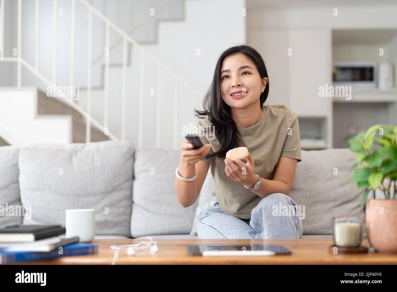 Immagine di una bella donna asiatica canale di ricerca con telecomando per guardare la tv mentre si siede sul divano a casa Foto Stock