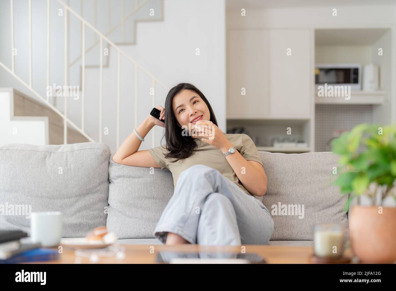 Immagine di una bella donna asiatica canale di ricerca con telecomando per guardare la tv mentre si siede sul divano a casa Foto Stock