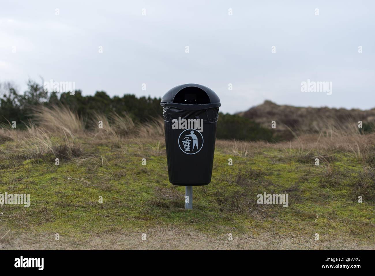 vuoto cestino nero con un sacco di rifiuti e pittogramma in piedi in erba nelle dune di sabbia in un giorno nuvoloso inverno Foto Stock