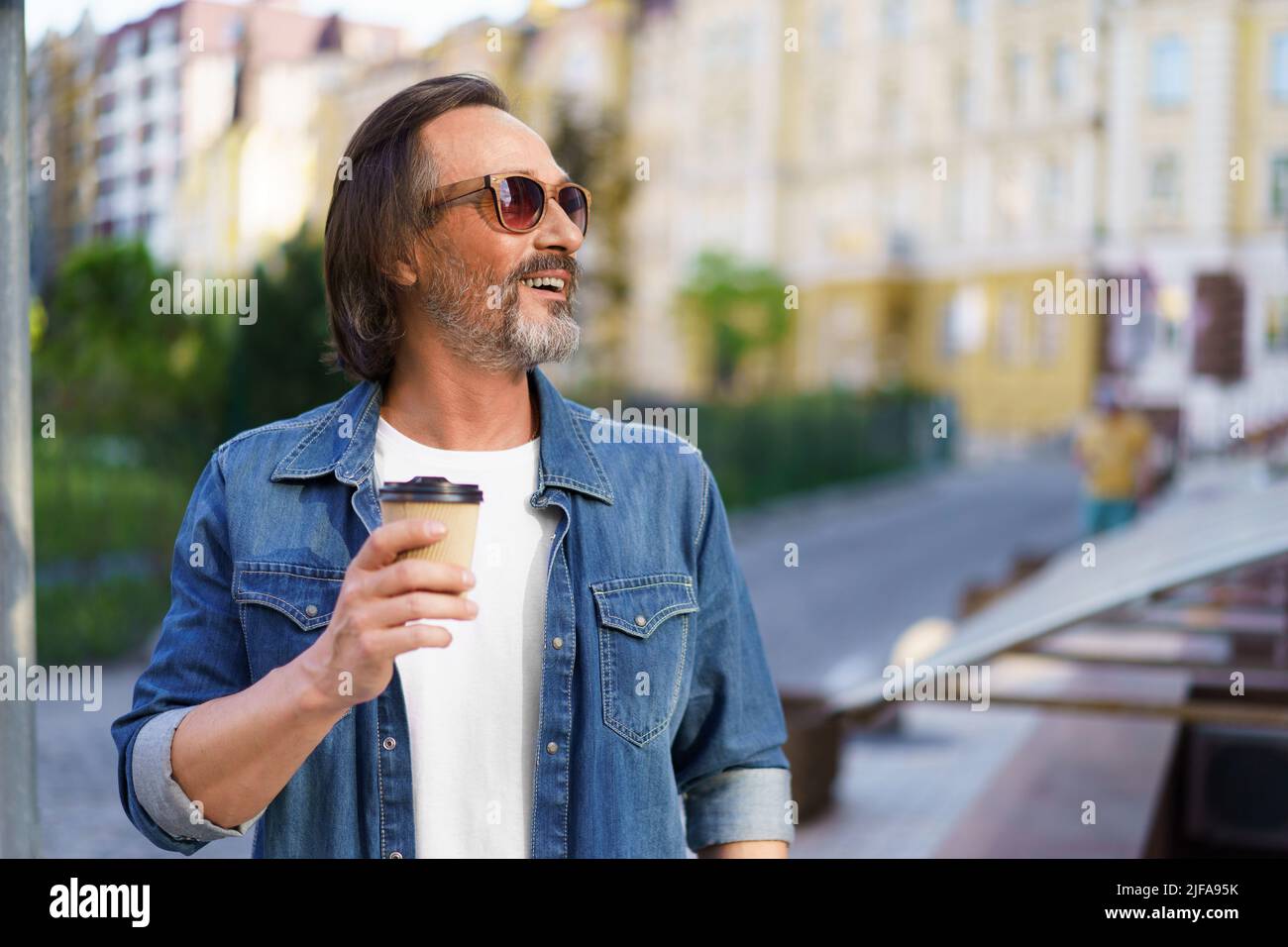 Uomo maturo sorridente guardando via lateralmente cattura ultimi raggi di sole indossare occhiali da sole con caffè in tazza di carta per le strade della città vecchia. Bell'uomo di mezza età con caffè take away. Foto Stock