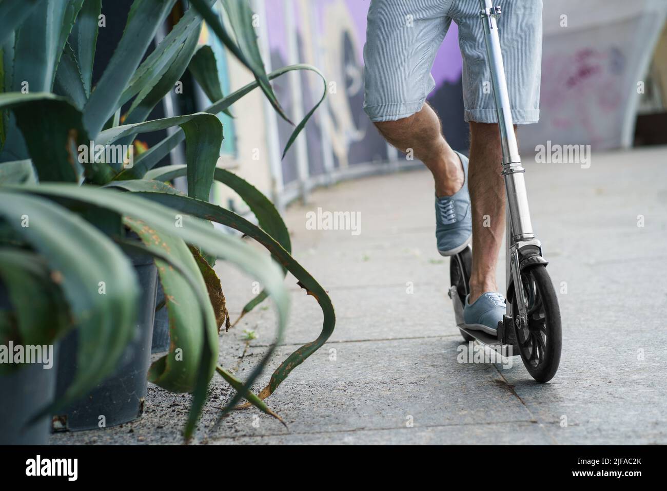Primo piano di un uomo che guida su uno scooter per adulti in città con ruote grandi in un elegante sneakers per le strade o parco con la spada lunga come agave pianta dopo il lavoro all'aperto. Nessuna faccia visibile. Concentrarsi sulla ruota. Foto Stock