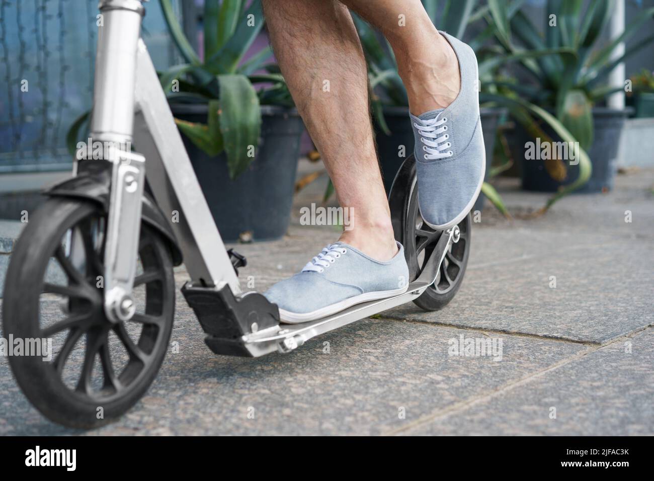 I piedi maschili in una sneakers si levano in piedi su uno scooter adulto della città con le ruote grandi che hanno un giro sulle strade o parcheggiano dopo il lavoro all'aperto con le piante di agave sullo sfondo. Nessuna faccia visibile. Mettere a fuoco la gamba sinistra. Foto Stock