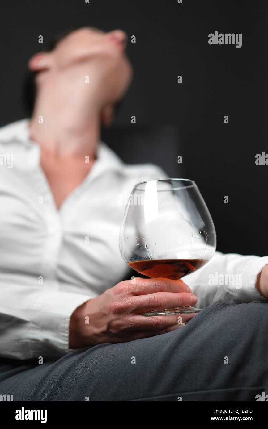 Bicchiere pieno di alcol tenuto da donna in abiti da lavoro seduti con le gambe incrociate e testa inclinata all'indietro . Concetto di depressione, Foto Stock