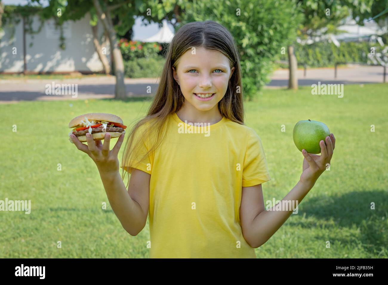 Una ragazza adolescente sceglie tra un hamburger e una mela all'aperto nel parco. Foto Stock